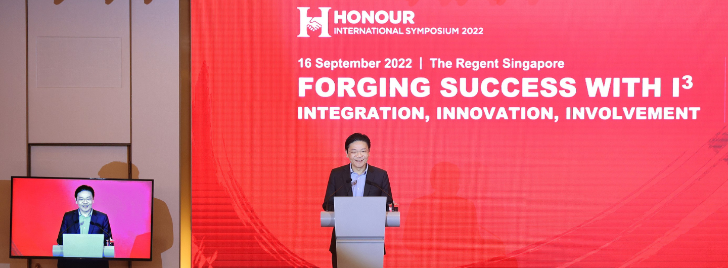 Man at podium, "Honour International Symposium 2022" red background, speech on monitor.